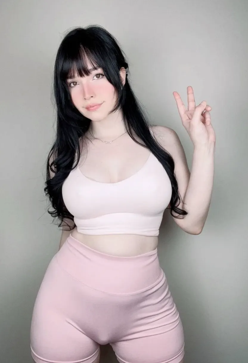 A young woman with long black hair and bangs posing against a plain background in a light pink sports bra and matching high-waisted shorts.