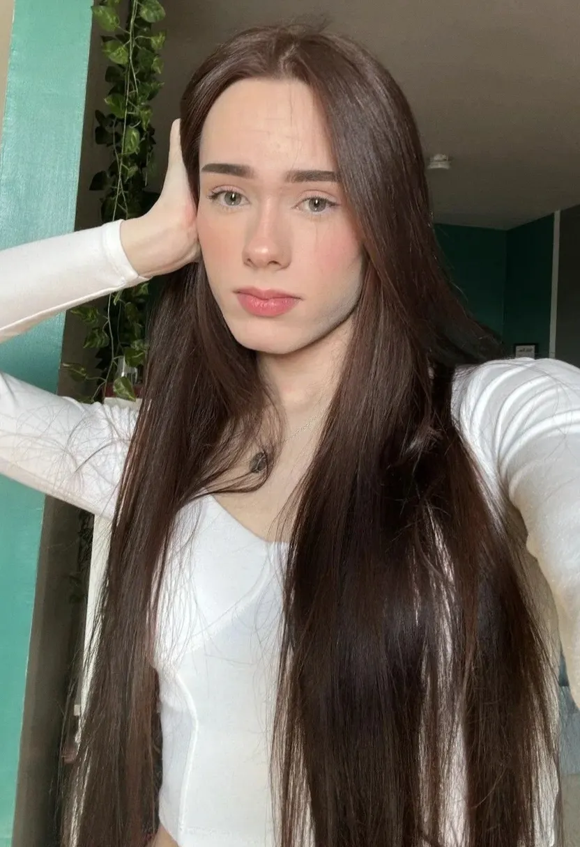 Young woman with long straight brown hair taking a close-up selfie in a white long-sleeve top.