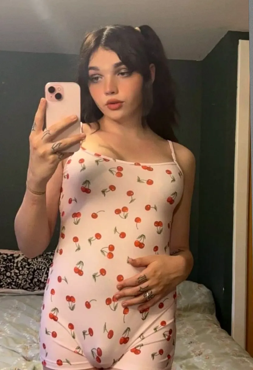 A young woman with dark hair in pigtails taking a mirror selfie in a bedroom with dark green walls, wearing a sleeveless cherry-patterned romper.