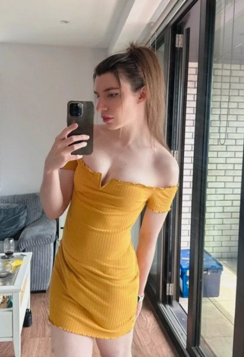 Woman with light brown hair pulled back taking a mirror selfie in a mustard yellow dress.