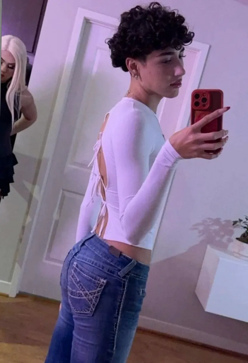 Person with short curly black hair taking a mirror selfie in a white top and blue jeans.