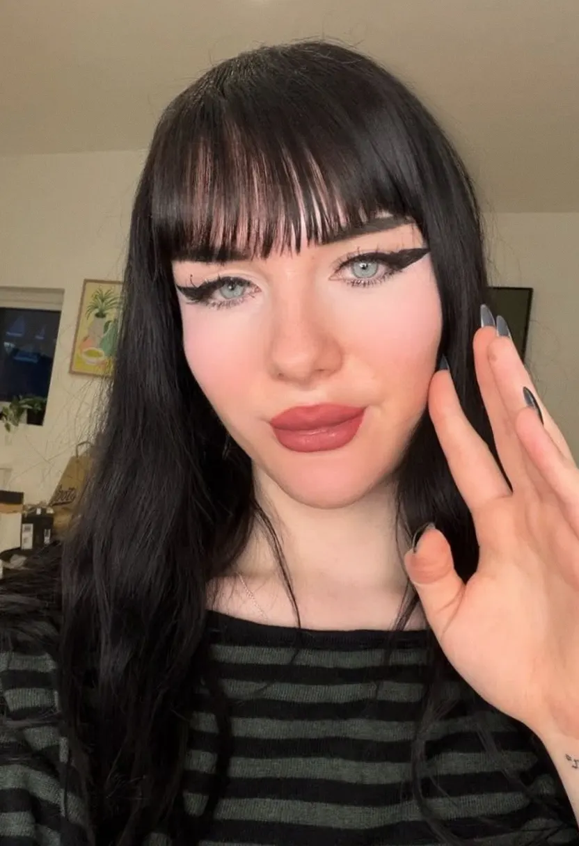 A young woman with long black hair and blunt bangs taking a close-up indoor selfie in a dark green and black striped top, with detailed makeup and a hand tattoo.